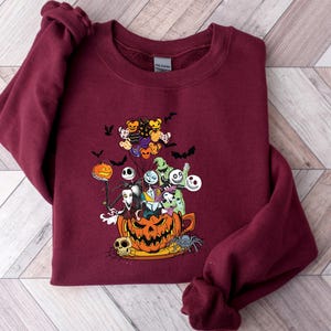 K&ouml;nnte beinhalten: Burgunderfarbenes Sweatshirt mit einer farbenfrohen Grafik von Charakteren aus The Nightmare Before Christmas. Das Design umfasst Jack Skellington, Sally, Zero und andere Charaktere in einem geschnitzten K&uuml;rbis. Flederm&auml;use und Luftballons vervollst&auml;ndigen das Halloween-Thema.