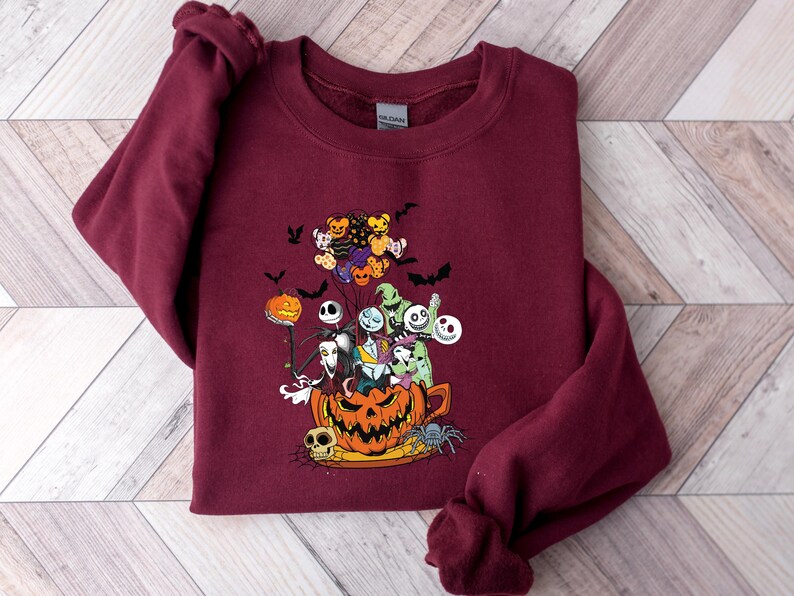 K&ouml;nnte beinhalten: Burgunderfarbenes Sweatshirt mit einer farbenfrohen Grafik von Charakteren aus The Nightmare Before Christmas. Das Design umfasst Jack Skellington, Sally, Zero und andere Charaktere in einem geschnitzten K&uuml;rbis. Flederm&auml;use und Luftballons vervollst&auml;ndigen das Halloween-Thema.