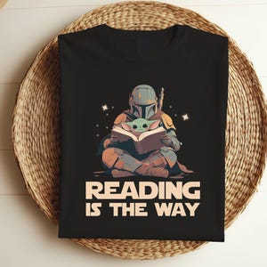 May include: Black t-shirt featuring a graphic of a Mandalorian character and Grogu reading a book. The text "READING IS THE WAY" is printed in white and tan. The shirt is folded on a woven basket.