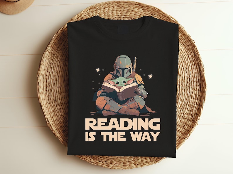 May include: Black t-shirt featuring a graphic of a Mandalorian character and Grogu reading a book. The text "READING IS THE WAY" is printed in white and tan. The shirt is folded on a woven basket.