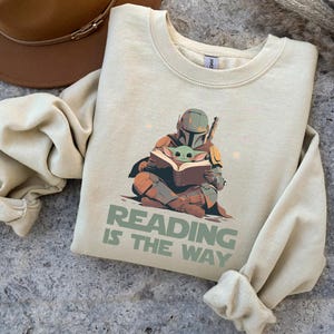 May include: Cream-colored sweatshirt featuring a graphic of the Mandalorian and Grogu reading a book. The text "READING IS THE WAY" is printed below the illustration. A brown hat is visible in the upper left corner.