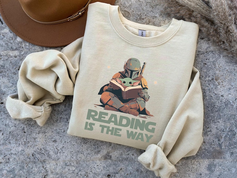 May include: Cream-colored sweatshirt featuring a graphic of the Mandalorian and Grogu reading a book. The text "READING IS THE WAY" is printed below the illustration. A brown hat is visible in the upper left corner.
