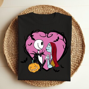 May include: Black t-shirt with a design of Jack Skellington and Sally from The Nightmare Before Christmas, framed by a pink heart and bats. A jack-o'-lantern is also included. The design has a romantic and spooky theme.