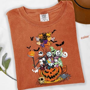 May include: Orange Comfort Colors t-shirt featuring a colorful graphic of characters from The Nightmare Before Christmas. The design includes Jack Skellington, Sally, and other characters, along with bats, pumpkins, and a spider. The text "color yam" is visible.