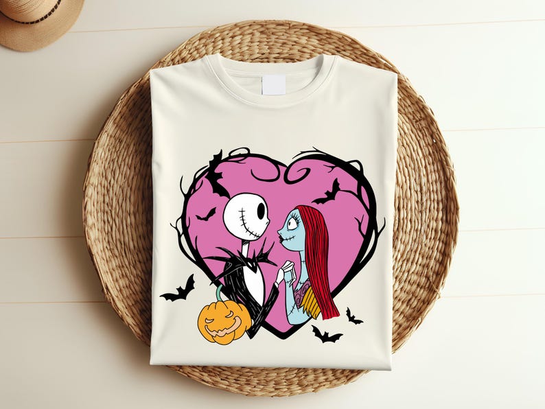 May include: Off-white t-shirt with a graphic of Jack Skellington and Sally from The Nightmare Before Christmas, within a pink heart-shaped frame with bats. A jack-o'-lantern is at the bottom. The design is on a woven basket.