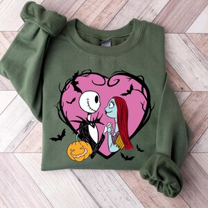 May include: An olive green sweatshirt with a graphic of Jack Skellington and Sally from The Nightmare Before Christmas, set within a pink heart with bats. A carved pumpkin is also included. The sweatshirt is folded on a light wood grain surface.