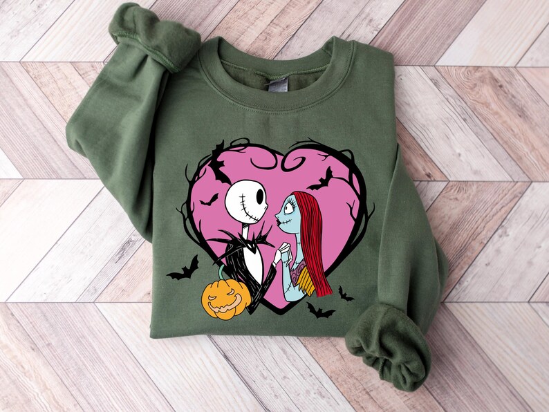 May include: An olive green sweatshirt with a graphic of Jack Skellington and Sally from The Nightmare Before Christmas, set within a pink heart with bats. A carved pumpkin is also included. The sweatshirt is folded on a light wood grain surface.