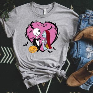 May include: A grey t-shirt with a graphic of Jack Skellington and Sally from The Nightmare Before Christmas, within a pink heart with bats. A pumpkin is also included in the design. The shirt is paired with jeans and boots.