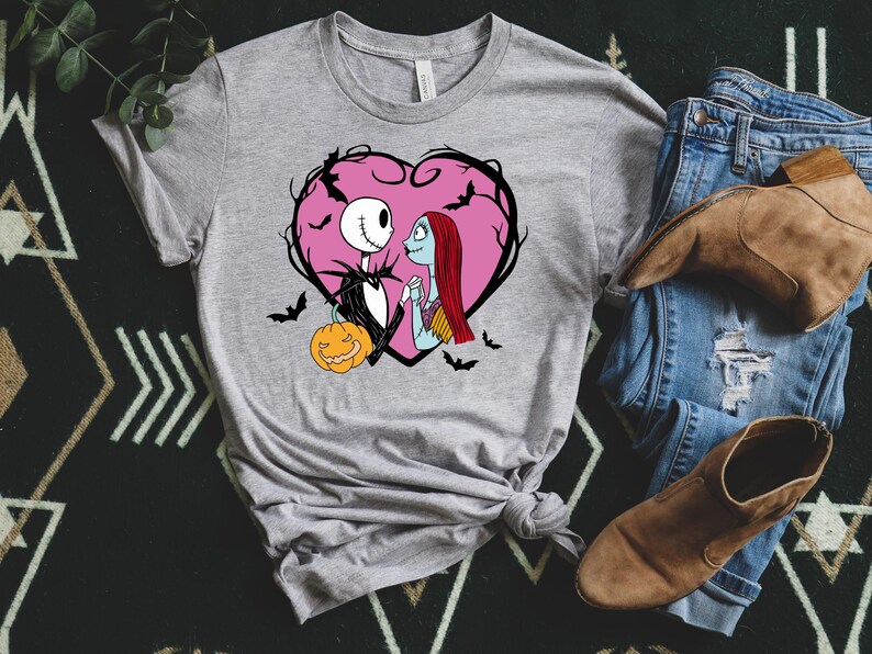 May include: A grey t-shirt with a graphic of Jack Skellington and Sally from The Nightmare Before Christmas, within a pink heart with bats. A pumpkin is also included in the design. The shirt is paired with jeans and boots.