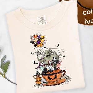 Pode incluir: Camiseta de cor marfim com um gr&aacute;fico tem&aacute;tico de Halloween. O design inclui personagens como Oogie Boogie e Jack Skellington, juntamente com bal&otilde;es, morcegos e uma x&iacute;cara de ch&aacute; em forma de ab&oacute;bora. A camisa tem uma etiqueta Comfort Colors.