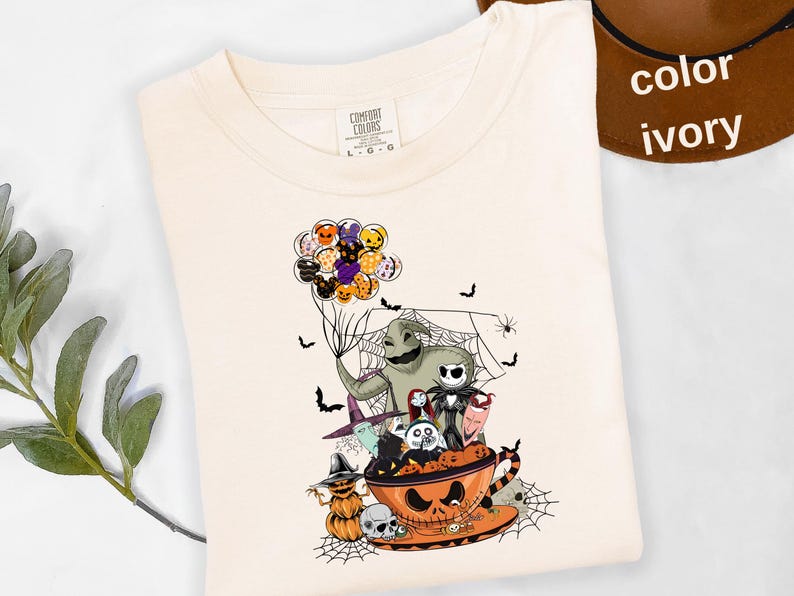 Pode incluir: Camiseta de cor marfim com um gr&aacute;fico tem&aacute;tico de Halloween. O design inclui personagens como Oogie Boogie e Jack Skellington, juntamente com bal&otilde;es, morcegos e uma x&iacute;cara de ch&aacute; em forma de ab&oacute;bora. A camisa tem uma etiqueta Comfort Colors.