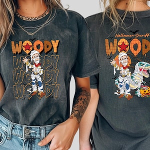 May include: Dark gray t-shirt featuring a Halloween-themed design. The front displays the word "WOODY" in orange and brown, with a cartoon character dressed as a mummy. The back says "Halloween Sheriff" with a similar design.