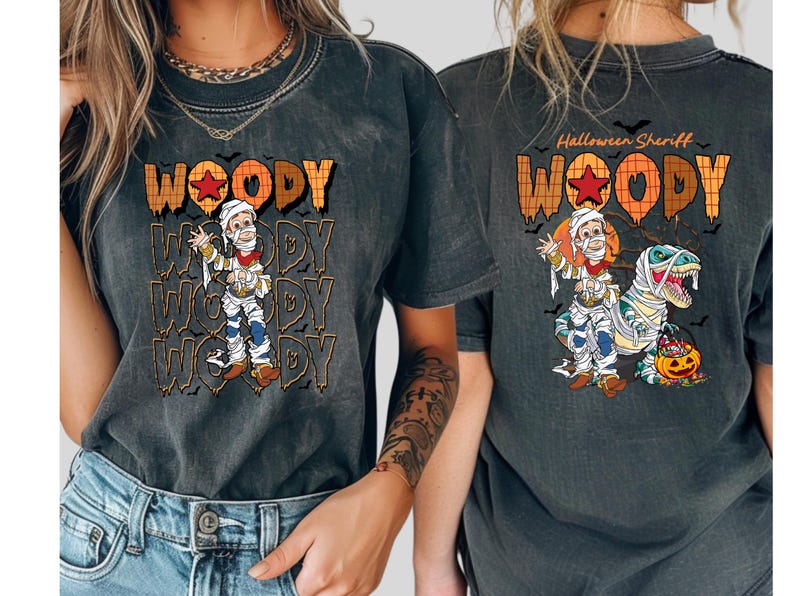 May include: Dark gray t-shirt featuring a Halloween-themed design. The front displays the word "WOODY" in orange and brown, with a cartoon character dressed as a mummy. The back says "Halloween Sheriff" with a similar design.