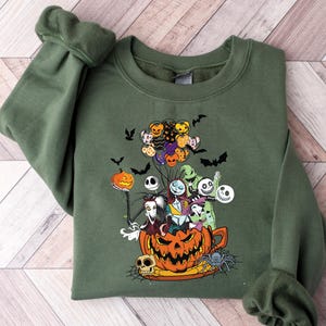 K&ouml;nnte beinhalten: Olivgr&uuml;nes Sweatshirt mit einem farbenfrohen Grafikdesign von Charakteren aus The Nightmare Before Christmas. Das Design zeigt Jack Skellington, Sally und andere Charaktere in einem K&uuml;rbis, mit Flederm&auml;usen und Luftballons. Ein gruseliges und festliches Design.
