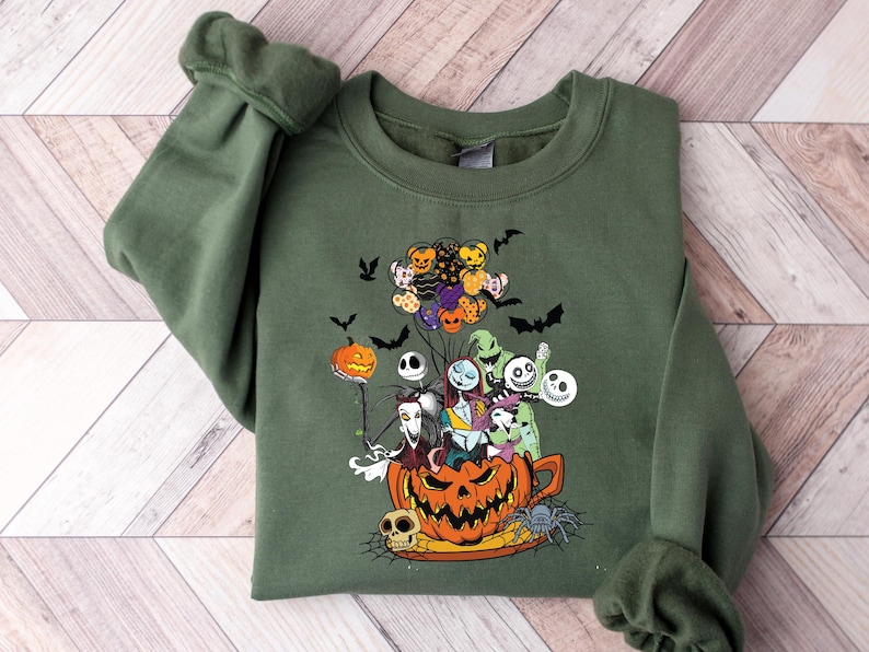 K&ouml;nnte beinhalten: Olivgr&uuml;nes Sweatshirt mit einem farbenfrohen Grafikdesign von Charakteren aus The Nightmare Before Christmas. Das Design zeigt Jack Skellington, Sally und andere Charaktere in einem K&uuml;rbis, mit Flederm&auml;usen und Luftballons. Ein gruseliges und festliches Design.