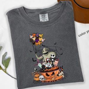 Pode incluir: Camiseta Comfort Colors cinza escuro com um gr&aacute;fico tem&aacute;tico de Halloween. O design inclui personagens como Oogie Boogie e Jack Skellington, e uma x&iacute;cara de ch&aacute; em forma de ab&oacute;bora. A camisa est&aacute; dobrada, com as palavras "color pepper" vis&iacute;veis.