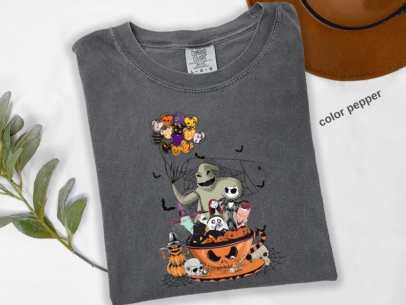 Pode incluir: Camiseta Comfort Colors cinza escuro com um gr&aacute;fico tem&aacute;tico de Halloween. O design inclui personagens como Oogie Boogie e Jack Skellington, e uma x&iacute;cara de ch&aacute; em forma de ab&oacute;bora. A camisa est&aacute; dobrada, com as palavras "color pepper" vis&iacute;veis.