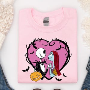 May include: A pale pink sweatshirt with a heart-shaped design featuring Jack Skellington and Sally from The Nightmare Before Christmas. The design includes a pumpkin and bats. The sweatshirt is a crew neck style.