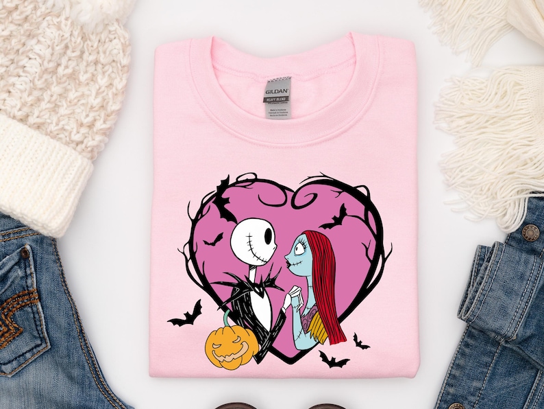 May include: A pale pink sweatshirt with a heart-shaped design featuring Jack Skellington and Sally from The Nightmare Before Christmas. The design includes a pumpkin and bats. The sweatshirt is a crew neck style.