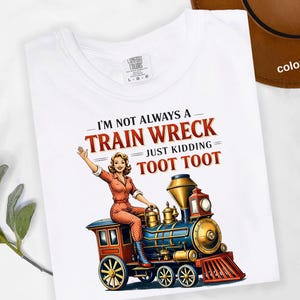 May include: White t-shirt with a colorful graphic of a woman on a train, with the text "I'M NOT ALWAYS A TRAIN WRECK JUST KIDDING = TOOT TOOT". The shirt has a crew neck and short sleeves.