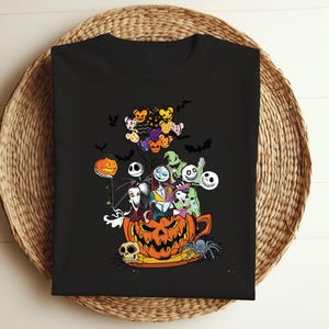 K&ouml;nnte beinhalten: Schwarzes T-Shirt mit einer farbenfrohen Grafik von Charakteren aus The Nightmare Before Christmas. Das Design umfasst Jack Skellington, Sally und andere Bewohner von Halloween Town sowie Flederm&auml;use und K&uuml;rbisse. Das T-Shirt wird auf einem geflochtenen Korb pr&auml;sentiert.