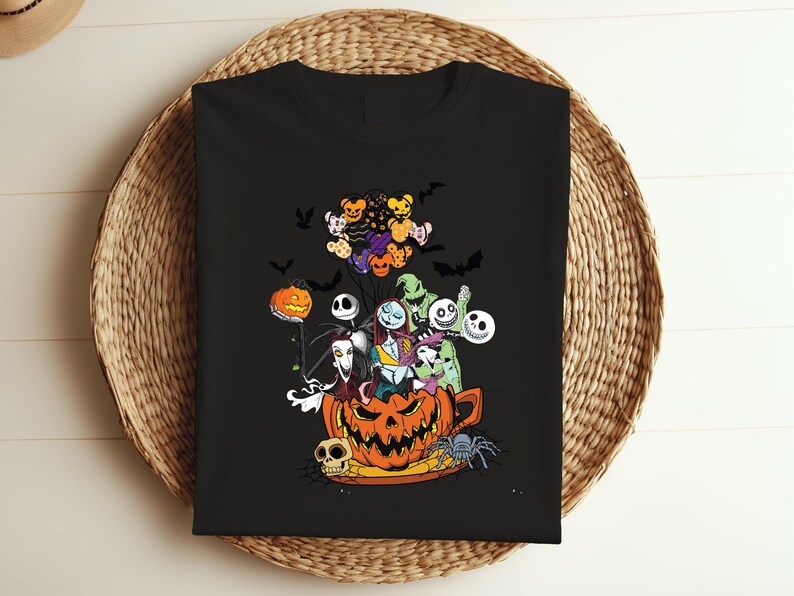 K&ouml;nnte beinhalten: Schwarzes T-Shirt mit einer farbenfrohen Grafik von Charakteren aus The Nightmare Before Christmas. Das Design umfasst Jack Skellington, Sally und andere Bewohner von Halloween Town sowie Flederm&auml;use und K&uuml;rbisse. Das T-Shirt wird auf einem geflochtenen Korb pr&auml;sentiert.