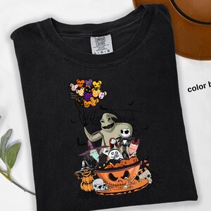 Pode incluir: Camiseta preta com um gr&aacute;fico tem&aacute;tico de Halloween. O design inclui personagens como Oogie Boogie e Jack Skellington, e uma tigela de ab&oacute;bora. Um grupo de bal&otilde;es de ursinhos de pel&uacute;cia flutua acima dos personagens. As palavras "color black" s&atilde;o vis&iacute;veis.