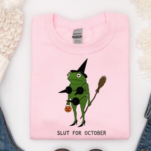 May include: Pink sweatshirt with a cartoon frog wearing a witch hat, bra, and heels, holding a broom and a jack-o'-lantern. The text "SLUT FOR OCTOBER" is printed below the frog. Halloween-themed apparel.