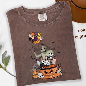 Pode incluir: Camiseta cor espresso com um gr&aacute;fico tem&aacute;tico de Halloween. O design inclui personagens como Oogie Boogie e Jack Skellington, junto com uma tigela de ab&oacute;bora e morcegos. A camisa tem uma etiqueta Comfort Colors.