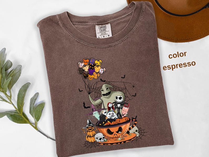Pode incluir: Camiseta cor espresso com um gr&aacute;fico tem&aacute;tico de Halloween. O design inclui personagens como Oogie Boogie e Jack Skellington, junto com uma tigela de ab&oacute;bora e morcegos. A camisa tem uma etiqueta Comfort Colors.