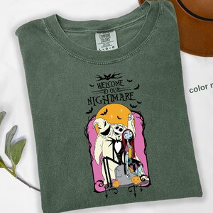 Puede incluir: Camiseta verde musgo con un dise&ntilde;o de "Pesadilla antes de Navidad". El dise&ntilde;o incluye a Jack Skellington, Sally y Oogie Boogie, con el texto "WELCOME TO OUR NIGHTMARE" y murci&eacute;lagos. Un sombrero marr&oacute;n es visible en el fondo.