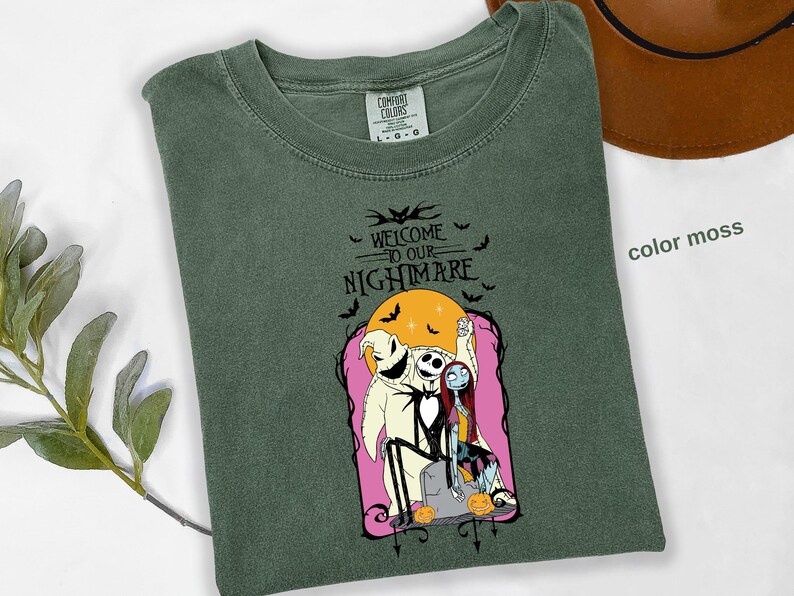 Puede incluir: Camiseta verde musgo con un dise&ntilde;o de "Pesadilla antes de Navidad". El dise&ntilde;o incluye a Jack Skellington, Sally y Oogie Boogie, con el texto "WELCOME TO OUR NIGHTMARE" y murci&eacute;lagos. Un sombrero marr&oacute;n es visible en el fondo.