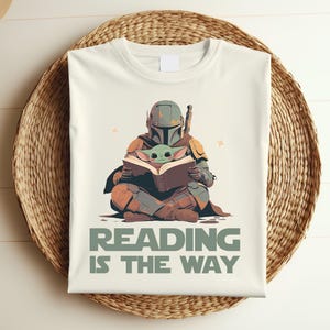 May include: Cream-colored t-shirt featuring a Mandalorian figure reading a book with Grogu. The design includes the text "READING IS THE WAY" in green. The shirt is displayed on a woven basket.