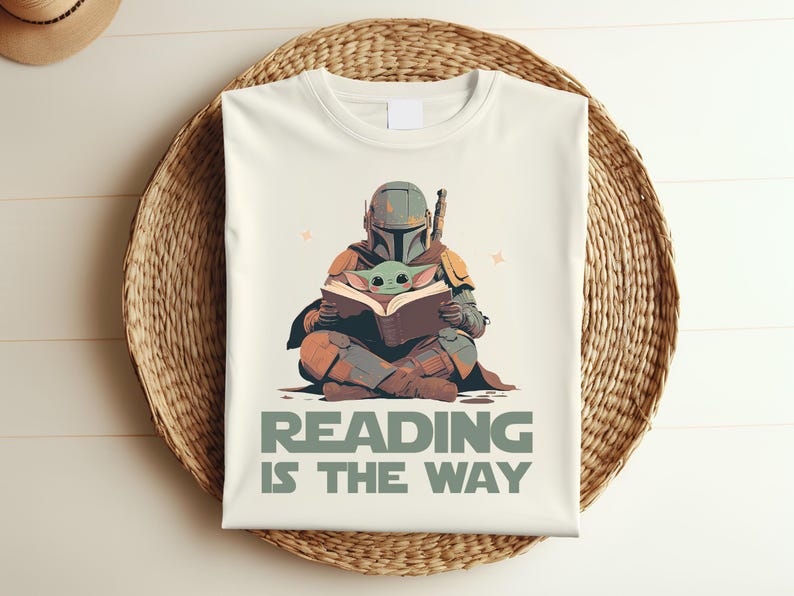 May include: Cream-colored t-shirt featuring a Mandalorian figure reading a book with Grogu. The design includes the text "READING IS THE WAY" in green. The shirt is displayed on a woven basket.