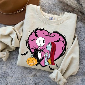 May include: Beige crew neck sweatshirt featuring a pink heart design with Jack Skellington and Sally from The Nightmare Before Christmas. The design includes bats and a jack-o'-lantern. A brown hat is visible in the background.