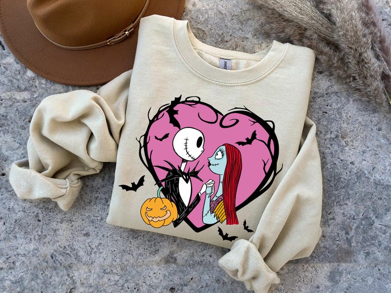 May include: Beige crew neck sweatshirt featuring a pink heart design with Jack Skellington and Sally from The Nightmare Before Christmas. The design includes bats and a jack-o'-lantern. A brown hat is visible in the background.