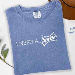 May include: Blue jean colored t-shirt with the text "I NEED A" and the Sprite logo in white. The shirt is folded and has a crew neck. A brown hat is partially visible in the background.