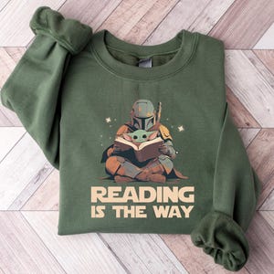 May include: Green sweatshirt featuring a graphic of a Mandalorian character and Grogu reading a book. The text "READING IS THE WAY" is printed below the illustration. The sweatshirt has long sleeves and a crew neck.