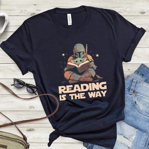 May include: Navy blue t-shirt featuring a graphic of a Mandalorian and Grogu reading a book, with the text "READING IS THE WAY". The design is in shades of brown, green, and orange. The shirt is paired with blue jeans and white sneakers.