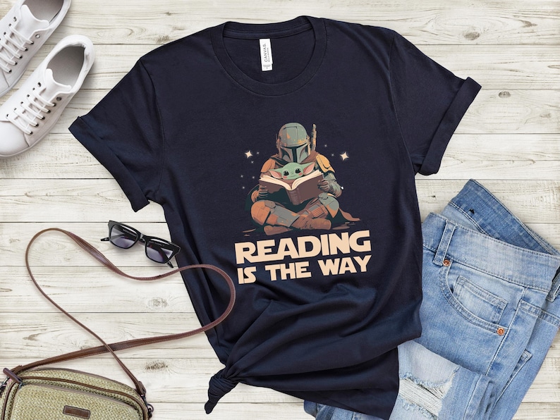 May include: Navy blue t-shirt featuring a graphic of a Mandalorian and Grogu reading a book, with the text "READING IS THE WAY". The design is in shades of brown, green, and orange. The shirt is paired with blue jeans and white sneakers.