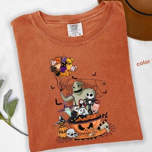 Pode incluir: Camiseta laranja Comfort Colors com um gr&aacute;fico tem&aacute;tico de Halloween. O design apresenta personagens de O Estranho Mundo de Jack, incluindo Jack Skellington e Oogie Boogie. A camisa tem gola redonda e mangas curtas.