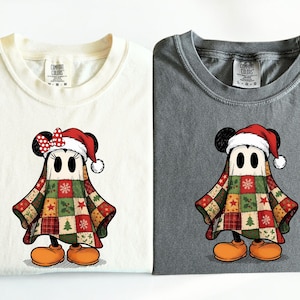 May include: Two folded t-shirts, one cream and one gray, each featuring a cartoon character dressed as a ghost for Christmas. The ghost wears a Santa hat and a patchwork quilt. The character has orange shoes.