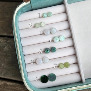 Op de afbeelding: Een witte juwelendoos met acht vakken, elk met een paar jade kralen. De kralen zijn genummerd van 1 tot 8. De kralen zijn in verschillende tinten groen en wit.