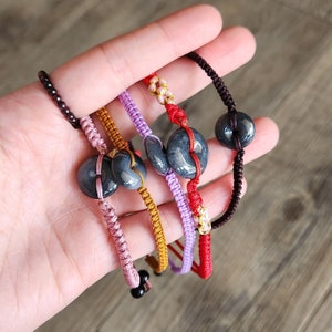 Puede incluir: Cinco pulseras con cuentas de jade negro y cuerdas de colores. Las pulseras están atadas juntas y están en una mano.