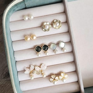 May include: A jewellery box with a light blue velvet lining and multiple compartments for storing earrings. The box is filled with various pearl and gemstone earrings, including flower, square, and butterfly designs.