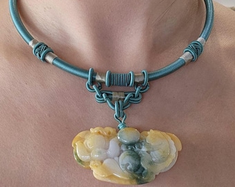 May include: A teal blue silk cord necklace with a silver and gold clasp. The necklace has a large jade pendant carved with a floral design in shades of green, yellow, and white.