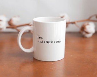 May include: White ceramic mug with a black inscription that reads "Tea. (n.) a hug in a cup."