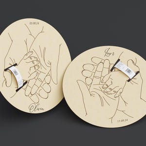 May include: Two light-colored wooden plaques with a hand-drawn illustration of hands holding a baby's hand. The plaques have the names "Olivia" and "Igor" with dates. Each plaque has a black bar with a white label.