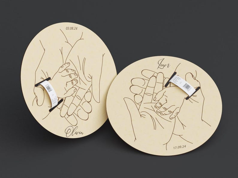May include: Two light-colored wooden plaques with a hand-drawn illustration of hands holding a baby's hand. The plaques have the names "Olivia" and "Igor" with dates. Each plaque has a black bar with a white label.