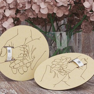 May include: Two light-colored wooden plaques with a hand-drawn illustration of hands holding a baby's hand. One plaque is round, the other is oval. A floral arrangement is in the background.